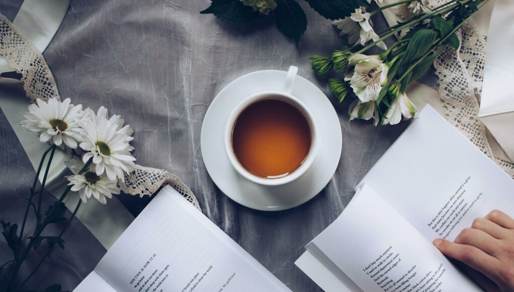 white ceramic teacup with saucer near two books above gray floral textile